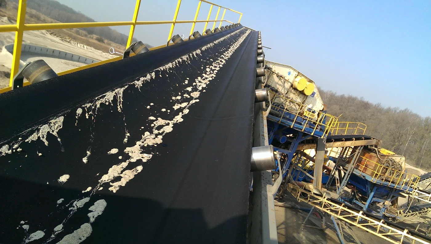 Großes Panoramabild eines langen, schwarzen Förderbandes, das sich diagonal durch die Mitte des Bildes erstreckt, von oben gesehen. Das Förderband ist leicht mit hellen, unregelmäßigen Sandflecken verschmutzt. Links vom Band, entlang seiner Kante, befindet sich eine gelbe Metallschutzbarriere mit Handlauf, die nach oben und rechts verläuft. Rechts vom Band sind Metallstützrollen sichtbar. Im Hintergrund, rechts vom Förderband, befindet sich eine große, komplexe Industriemaschinenstruktur in Gelb und Blau, die zum Zerkleinern von Materialien verwendet wird. In der Ferne rechts vom Förderband sind Bäume und ein Fragment eines Waldes sichtbar, während links vom Horizont ein offenes Bergbaugebiet mit sichtbaren Sand- und Schutthaufen zu sehen ist. Die gesamte Szene wird von hellem Tageslicht beleuchtet, und der Himmel ist klar und blau.